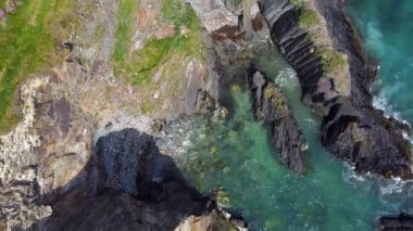 Turkuaz suyu olan kayalık bir sahil şeridinin havadan görüntüsü. Kayalar koyu gri ve dokulu. Beyaz kapaklar su yüzeyinde görülebilir..