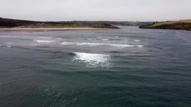 Kumlu bir sahil ve mavi okyanus, beyaz binaları olan yeşil bir tepe tarafından çerçevelenmiştir. Su derin bir mavidir ve kıyıda birkaç küçük dalga kırılır..