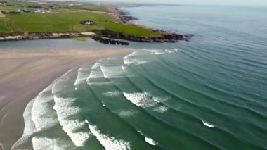 Yukarıdan çekilen kayalık bir kıyı şeridinin panoramik görüntüsü. Okyanus derin bir mavidir, dalgalar gelir ve kıyıya vurur. Uzaktaki çimenli bir uçurumun üzerinde küçük bir ev oturuyor..