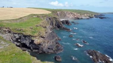Üzerinde yeşil bir tepe olan kayalık bir uçurum manzarası. Uçurumun önünde açık mavi bir deniz ve yukarıda mavi bir gökyüzü var..