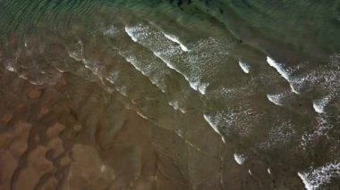 Dalgalı ve kumlu bir su kütlesi. Su sakin gibi görünüyor. Sahne huzurlu ve sakin, dalgalar yavaşça kıyıya vururken.