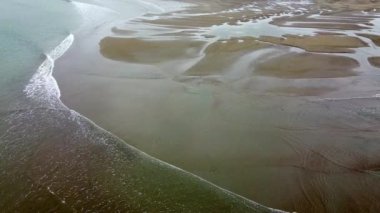 Yukarıdan çekilen bir plaj sahnesi, çekilen gelgiti gösteriyor. Kumlu sahil, geri çekilen dalgaların oluşturduğu sığ su havuzları ve kumsallarla işaretlenmiştir..