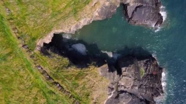Kayalık bir kıyı şeridinin yukarıdan görünüşü. Koyu gri kayalar, turkuaz su ve beyaz kapaklarla keskin bir tezat oluşturuyor..