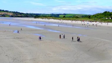 West Cork, İrlanda, 20 Ağustos 2024. Kumsalda birkaç kişi kumsalda yürüyor. Dalgalar alçak, yüzeysel su birikintileri ortaya çıkıyor. Uzakta yeşil tepeler ve açık mavi bir gökyüzü.