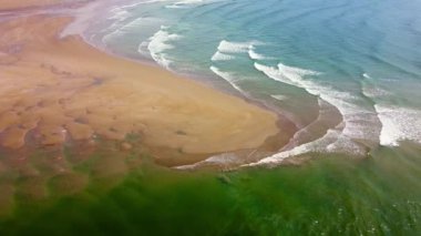 Dalgalar, alçak gelgit sırasında kumlu bir sahilde suların ve kıyı şeridi boyunca karanın bir karışımını gösteren desenler oluşturur..