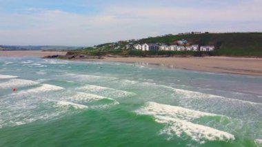 Okyanus derin bir mavidir, kıyıda beyaz dalgalar kırılır. Kumsal kumlu ve arkasında beyaz binalar olan yeşil bir tepe var. Gökyüzü açık mavi ve birkaç ince bulut var..