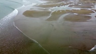 Suyun çekildiği kumlu bir sahilin havadan görünüşü kumsalların ve kanalların karmaşık desenlerini ortaya çıkarıyor. Su sakin ve sığ, açık mavi bir tonda gökyüzünü yansıtıyor..