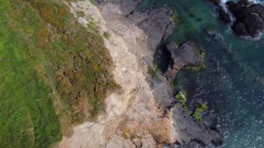 Kayalık bir kıyı şeridinin yukarıdan görünüşü. Koyu gri kayalar turkuaz su ve beyaz kapaklarla keskin bir tezat oluşturuyor. Su, deniz tabanını görmek için yeterince berrak. Kum ve yosun karışımı ortaya çıkıyor..