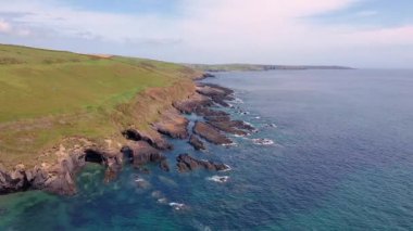Üzerinde yeşil çimenli bir tepe olan kayalık bir sahil şeridi. Su berrak mavi ve gökyüzü parçalı bulutlu..