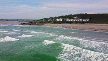 Uzun, kumlu bir sahil üzerinde beyaz binalar olan yeşil bir tepenin etrafını sarmıştır. Okyanus derin bir mavidir, kıyıda beyaz dalgalar kırılır. Gökyüzü açık mavi ve birkaç ince bulut var..