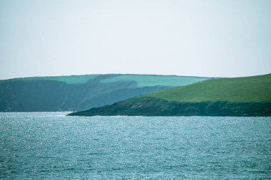 Berrak bir günde berrak bir su kütlesine doğru eğimli yemyeşil tepeleri gösteren sakin bir manzara, güneş ışığını yansıtıyor ve doğal güzelliği arttırıyor..