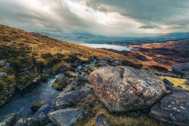 Kayalık arazinin nefes kesen manzarası bulutlarla dolu kasvetli bir gökyüzünün altında arka planda yuvarlanan tepelerle sakin bir göle doğru iniyor..