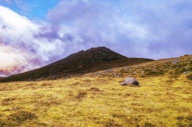 Çimenli bir yamaç, parçalı bulutlu bir gökyüzünün altındaki bir dağ zirvesine doğru yükseliyor. Dağılmış kayalar manzaranın ön planına nokta koyuyor..