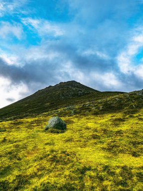Büyük, yuvarlak bir kaya çimenli bir tepenin önünde oturur. Tepe, parçalı bulutlu bir gökyüzünün altında bir dağ zirvesine doğru yükseliyor..