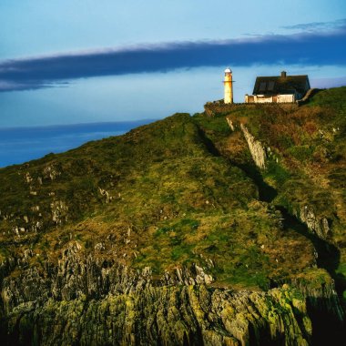 Beyaz bir deniz feneri kayalık bir uçurumun üzerinde, gri çatılı, küçük, beyaz, tek katlı bir evin yanında duruyor. Deniz feneri uzun ve ince, beyaz silindirik bir gövdesi ve siyah bir feneri var..
