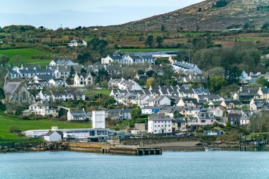 Yarımadanın kenarında bir ev kümesi olan bir kıyı manzarası. Evler çoğunlukla beyazdır ve siyah çatıları vardır. Evlerin önünde kayalık bir sahil ve bir su kütlesi var..