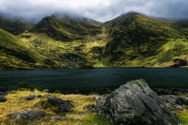 Kayalık bir kıyı yamaçları, iki büyük, yeşil dağlı, durgun, karanlık bir su gölüne kadar uzanır..