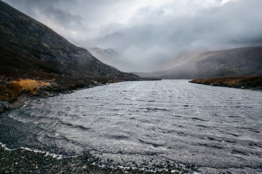 Yuvarlanan tepeleri ve merkezinde dolambaçlı gri bir nehri olan puslu, düşünceli bir manzara, sessiz bir yalnızlık ve içsel bir sükunet ortamı yaratır..