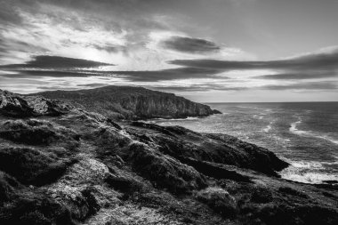 Günbatımında dramatik bir gökyüzü altında engebeli kayalıkları ve çalkantılı okyanusu vurgulayan çarpıcı bir siyah beyaz kıyı manzarası yer alıyor..