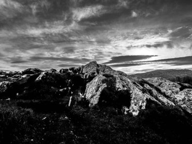 Engebeli bir arazide, alacakaranlıkta dramatik ve bulutlu bir gökyüzüne karşı dizilmiş büyük taşlar, siyah ve beyaz tonlarda çarpıcı bir kontrast oluşturur..