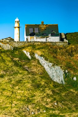 Kayalık bir uçurumun üzerinde beyaz bir deniz feneri duruyor. Gökyüzü açık mavi ve güneş parlıyor..