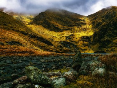 Küçük, durgun bir göl, ince bulutlarla kaplı iki dağ yamacı ve büyük, yuvarlak kayaların arasında yuva yapar..