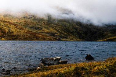 Sakin bir göl, sonbaharın sonlarında etrafındaki sisli tepeleri yansıtır. Topraksı renklerle ve dokularla dolu doğal bir ortamda huzurlu bir atmosfer yaratır..