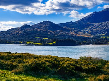 Sakin mavi bir su kütlesi, kayalık bir kıyı şeridi ve dağlardan oluşan bir manzara..