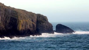 Büyük, koyu gri bir uçurum yüzeyi okyanustan yükseliyor, engebeli yüzeyi sarı ve yeşil çizgilerle kaplı. Dalgalar kayalık zemine çarpar ve çalkantılı bir sahne yaratır..