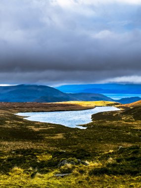Sarmal bir nehir yemyeşil tepelerin arasından akar, yukarıdaki dramatik gökyüzünü yansıtır. Sakin manzara, uzak bir bölgedeki doğanın güzelliğini gözler önüne seriyor..