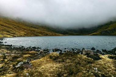 Bir dağ gölünün sakin yüzeyi yükselen sisi yansıtırken, yemyeşil tepeler arka planda beliriyor, şafak vakti sakin bir doğal manzara yaratıyor..