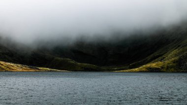 Sakin bir göl, yoğun bulutların altında sessizce oturur ve arka planda yeşil tepeler yükselir. Atmosfer sakin. Huzur ve macera hissi uyandırıyor..