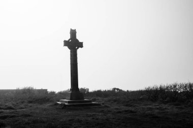 A tall, dark cross is positioned on a grassy hilltop, surrounded by sparse vegetation and a misty skyline. The atmosphere feels serene and reflective during the late afternoon. clipart