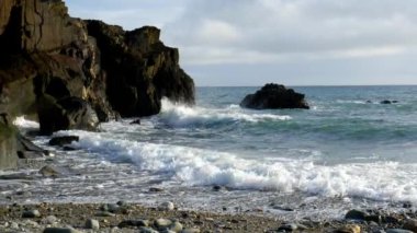 Çalkantılı bir denize bakan kayalık bir tepe. Dalgalar kıyıya çarpıyor ve beyaz köpük oluşturuyor. Gökyüzü soluk bir mavi ve bir kaç ince bulut.
