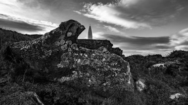 Uzakta uzun, beyaz, dar bir yapı ve çimen parçalarıyla kayalık bir yamaç manzarası. Gökyüzü çoğunlukla açık..