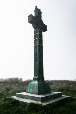 Sis kaplı alanlardan yükselen uzun bir Kelt haçı heykeli karmaşık oymalar ve desenler sergiliyor, dingin İrlanda manzarasındaki miras ve maneviyatı sembolize ediyor.