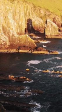 Dik, kayalık kayalıkları ve karanlık suyu gösteren kıyı manzarası. Daha küçük kayalar ve dalgalar görülebilir. Uçurumlar kısmen çimlerle kaplı..