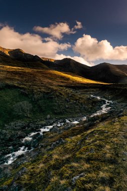 Akşamın erken saatlerinde bulutlu bir gökyüzünün altında akan bir dereyle dağlık bir arazi..