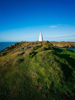 A historic lighthouse rises above the green hills overlooking the Atlantic Ocean. Bright blue skies add to the tranquil atmosphere of this coastal landscape. clipart