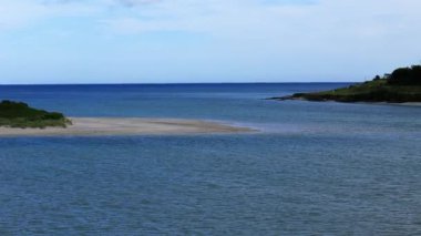 Açık mavi suların ortasında parlak bir gökyüzünün altında kumlu bir sahil belirir. Huzurlu atmosfer, kıyıdaki rahatlama ve tefekküre davetiye çıkarır..