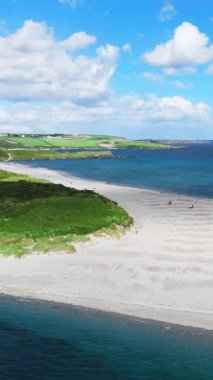 El değmemiş bir plajı gösteren yüksek açılı bir fotoğraf. Açık kum, gök mavisi suya zıt düşer. Yeşil sahil tepeleri görünür.