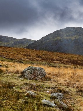 Engebeli bir arazide, uzak dağların üzerinde süzülen kara bulutlarla çevrili geniş kayalar bulunur ve dramatik bir atmosfer yaratır..