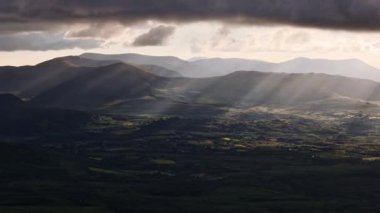Yuvarlanan tepeler ve vadiler kısmen bulutlu bir gökyüzünün altındaki uzak dağlara kadar uzanır. Güneş ışığı ışık ışınları oluşturarak filtrelenir.