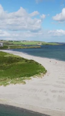 Plaj manzarası açık renkli bir kumsalın okyanusa doğru uzandığını gösteriyor. Yeşil arazi ve binalar uzakta..