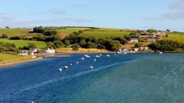 Sahil evleri sakin bir körfez hizasında. Birçok küçük tekne sakin sulara demirlemiştir. Yuvarlanan yeşil tepeler resim gibi bir zemin oluşturuyor..