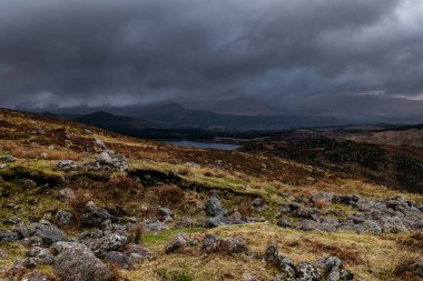 Kasvetli bir öğleden sonra engebeli kırsal bölgede, manzaralı dağların ve gölün üzerinde kara bulutlar süzülür..