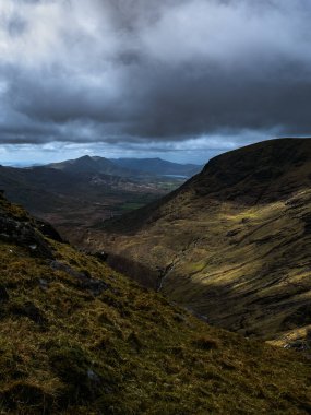Engebeli bir manzaranın yüksek açılı görüntüsü. Gölgeler ön plandaki yamaçlara hakim olurken, güneş ışığı uzak tepelere ve dolambaçlı bir akıntıya dokunuyor..