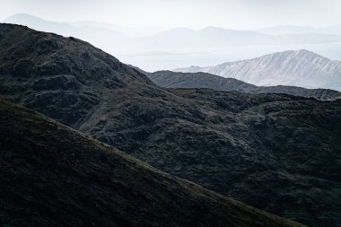 Karanlık, kayalık yamaçlar uzak, puslu bir su kütlesine doğru iniyor. Dağların katmanları solgun bir gökyüzünün altına uzanıyor..