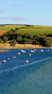 Huzurlu bir sahil manzarası. Beyaz evler sakin bir körfezin yanında kümelenmiş, küçük teknelerle benekli. Yeşil tepeler yavaşça arka planda yükselir.