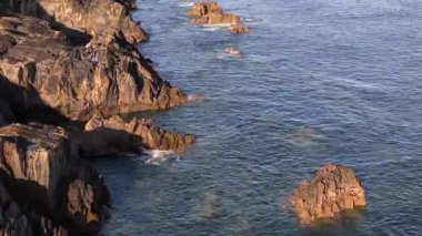 Güneş batarken dalgalar sivri kayalara çarpar ve sakin okyanusun üzerine sıcak bir parıltı saçar. Bu resim gibi manzara, sahil boyunca doğanın güzelliğini yansıtıyor..